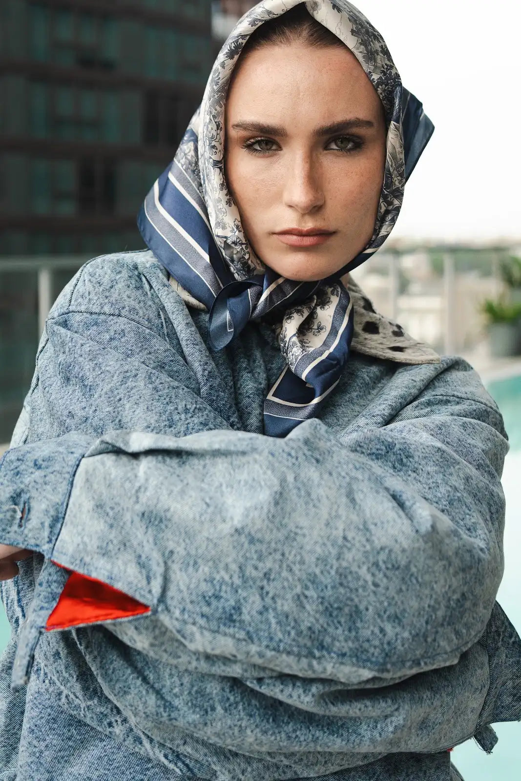 Model wearing a cropped blue denim jacket and printed navy scarf by the pool.