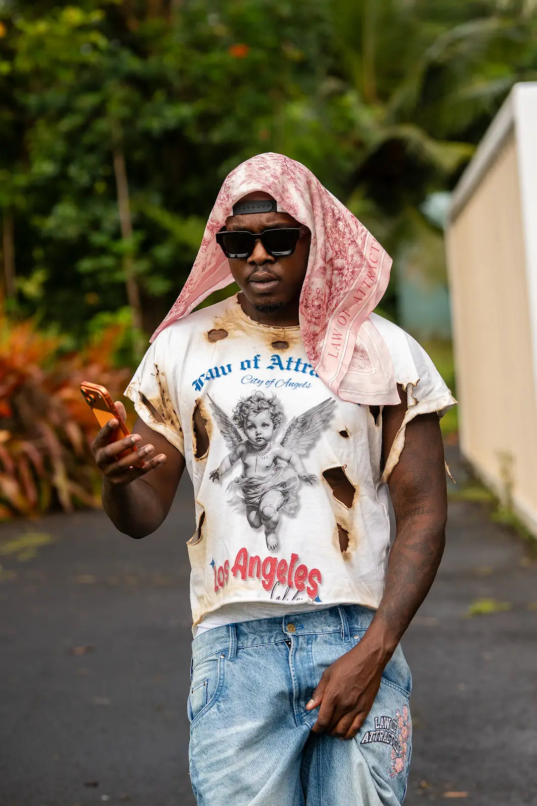 Model wearing a pink printed scarf over the head with sunglasses, a white City of Angels tee, and blue jeans.