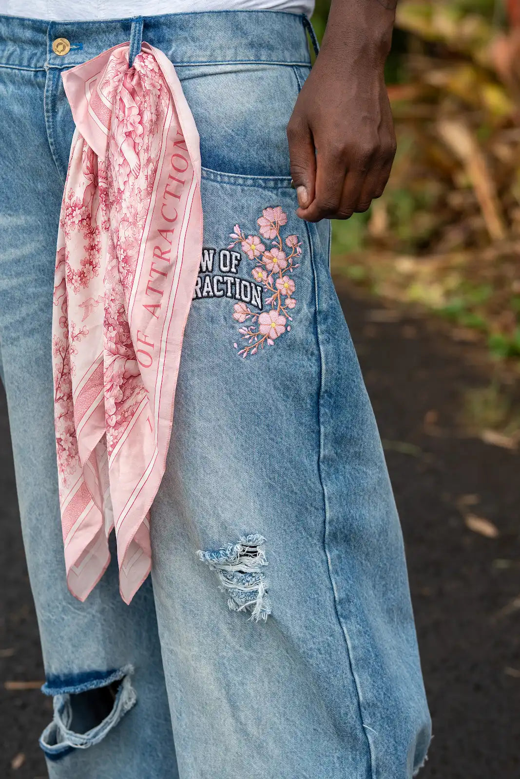 Close-up of a pink printed Law of Attraction scarf tied to a belt loop on floral-embroidered blue jeans.