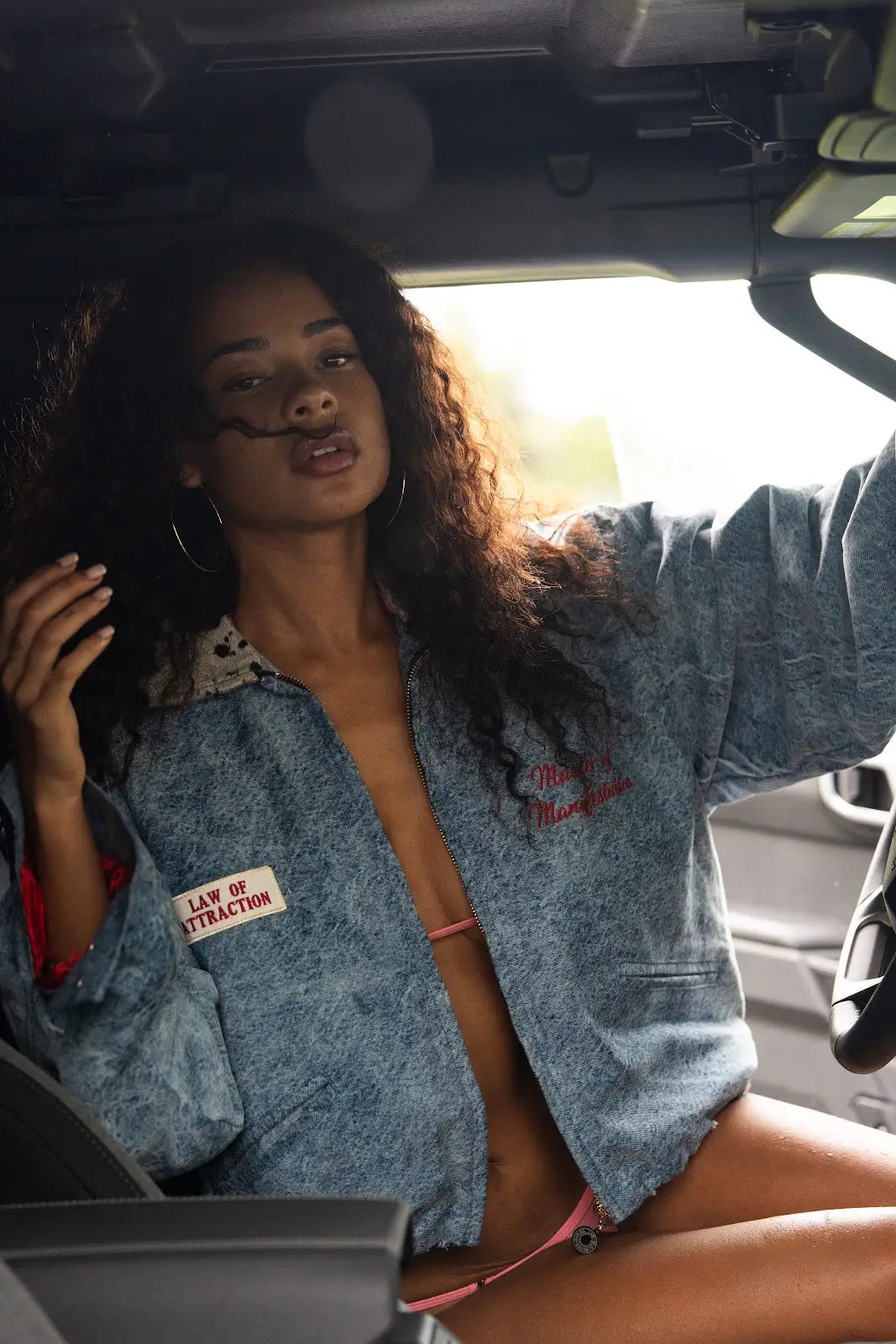 Model seated inside a vehicle wearing a cropped blue denim jacket with a chest label and embroidered lettering.