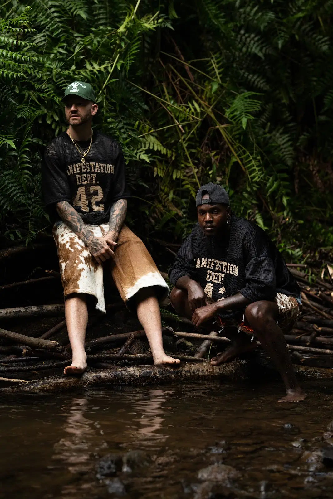 Two models wearing black 42 jerseys and brown cowhide shorts beside a tropical stream.