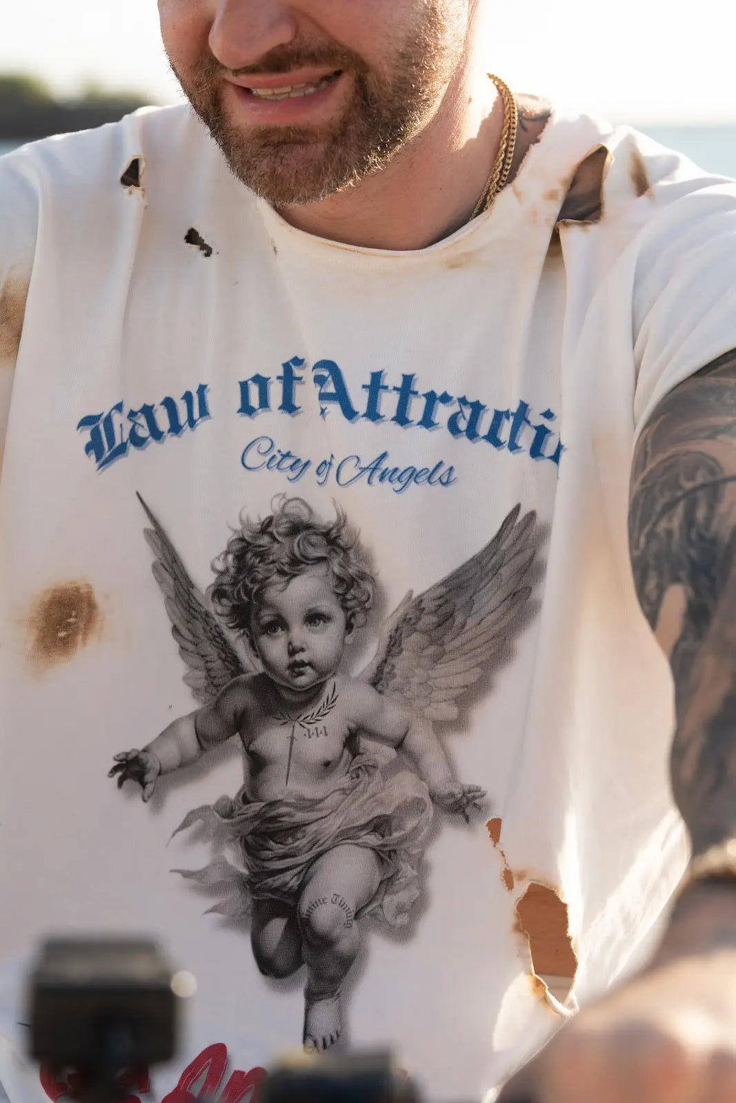 Close-up of a white Law of Attraction City of Angels tee with blue lettering and a black cherub graphic.