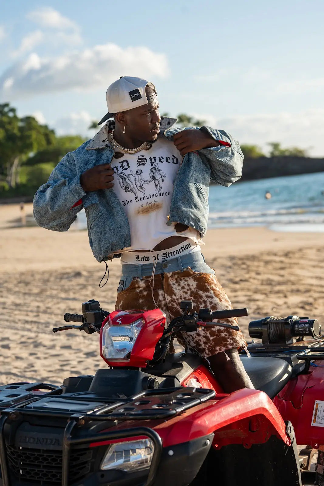 Model wearing a cropped blue denim jacket over a white God Speed graphic tee and brown cowhide shorts beside a red quad bike on the beach.