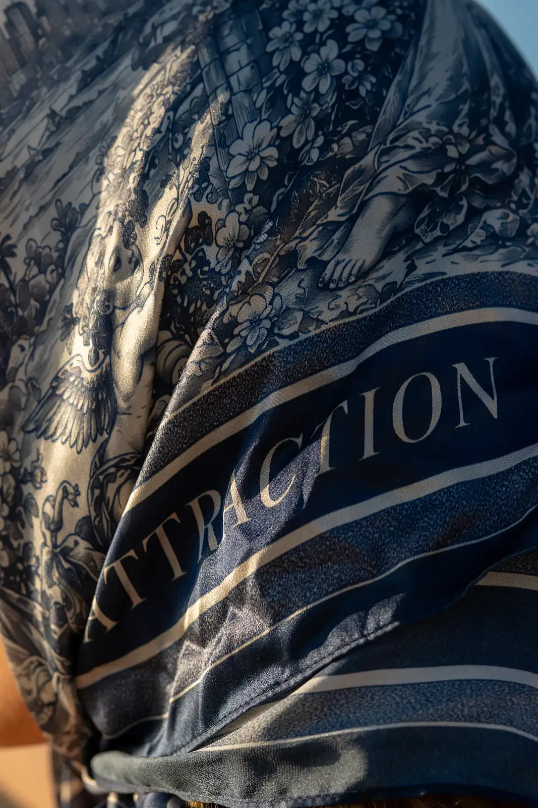 Detail of the navy scarf's floral and figurative print, flowing folds, border, and Attraction lettering.