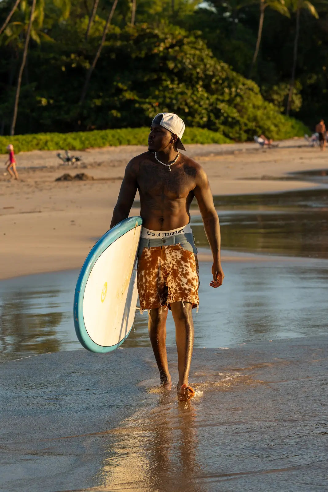Model walking along the beach in brown-and-white cowhide shorts while carrying a surfboard.