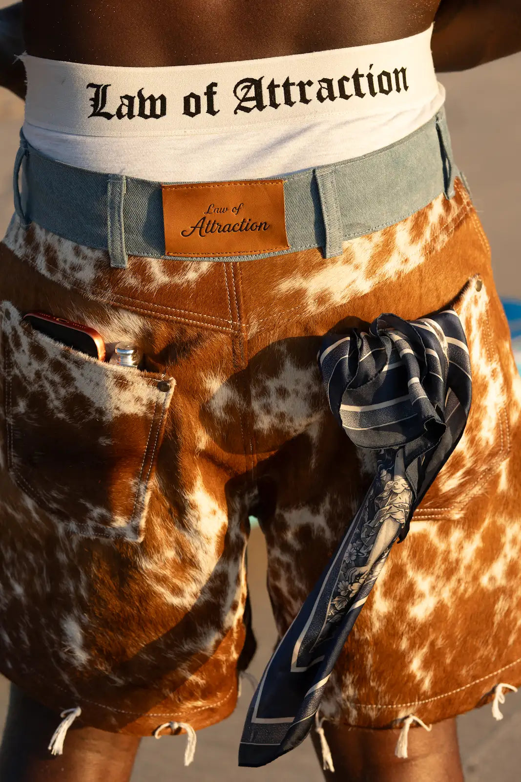 Detail of the brown cowhide shorts' blue denim waistband, tan leather Law of Attraction patch, back pocket, and navy scarf.