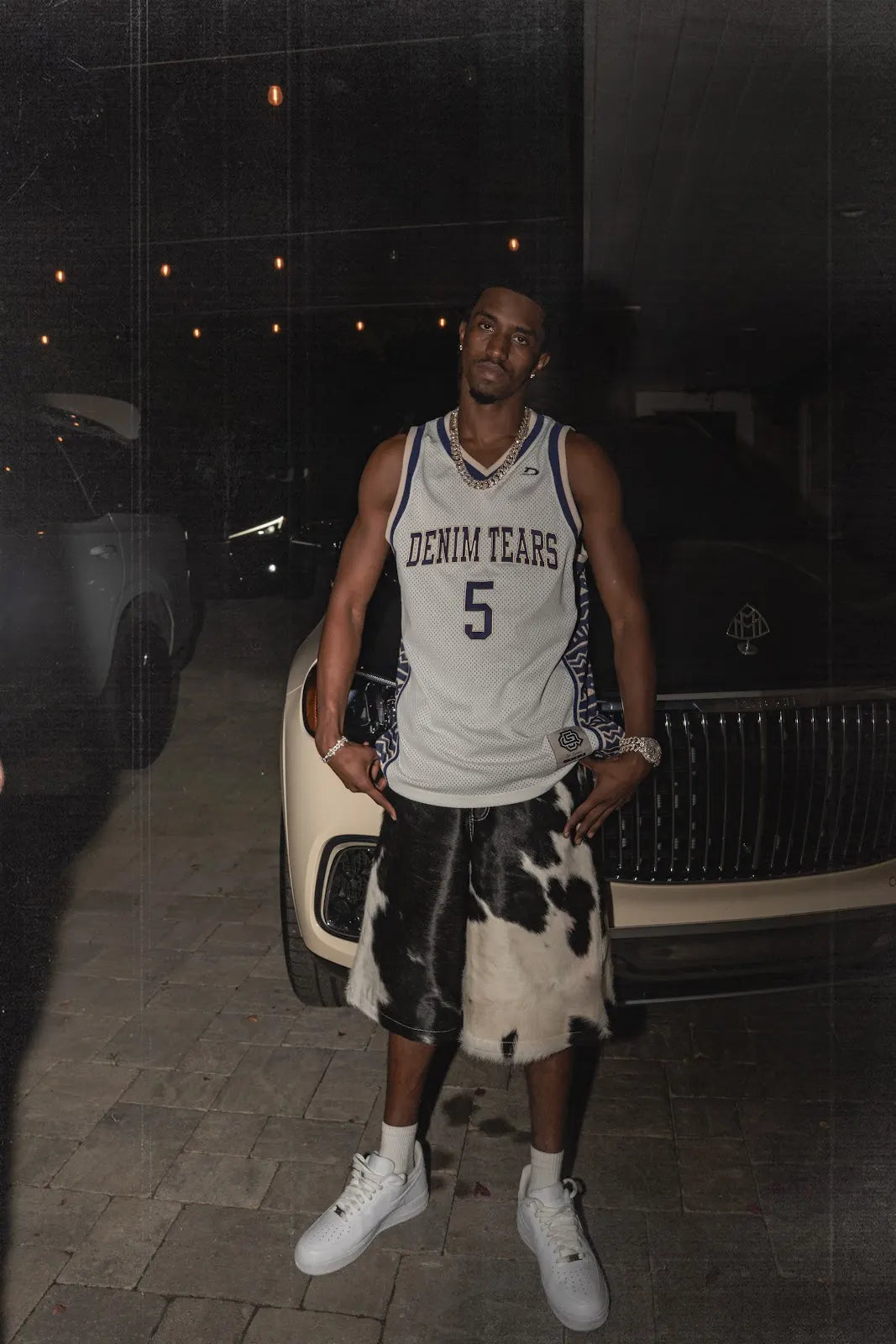 Christian Combs standing beside a parked car in black-and-white cowhide jorts, a white basketball jersey and white sneakers.