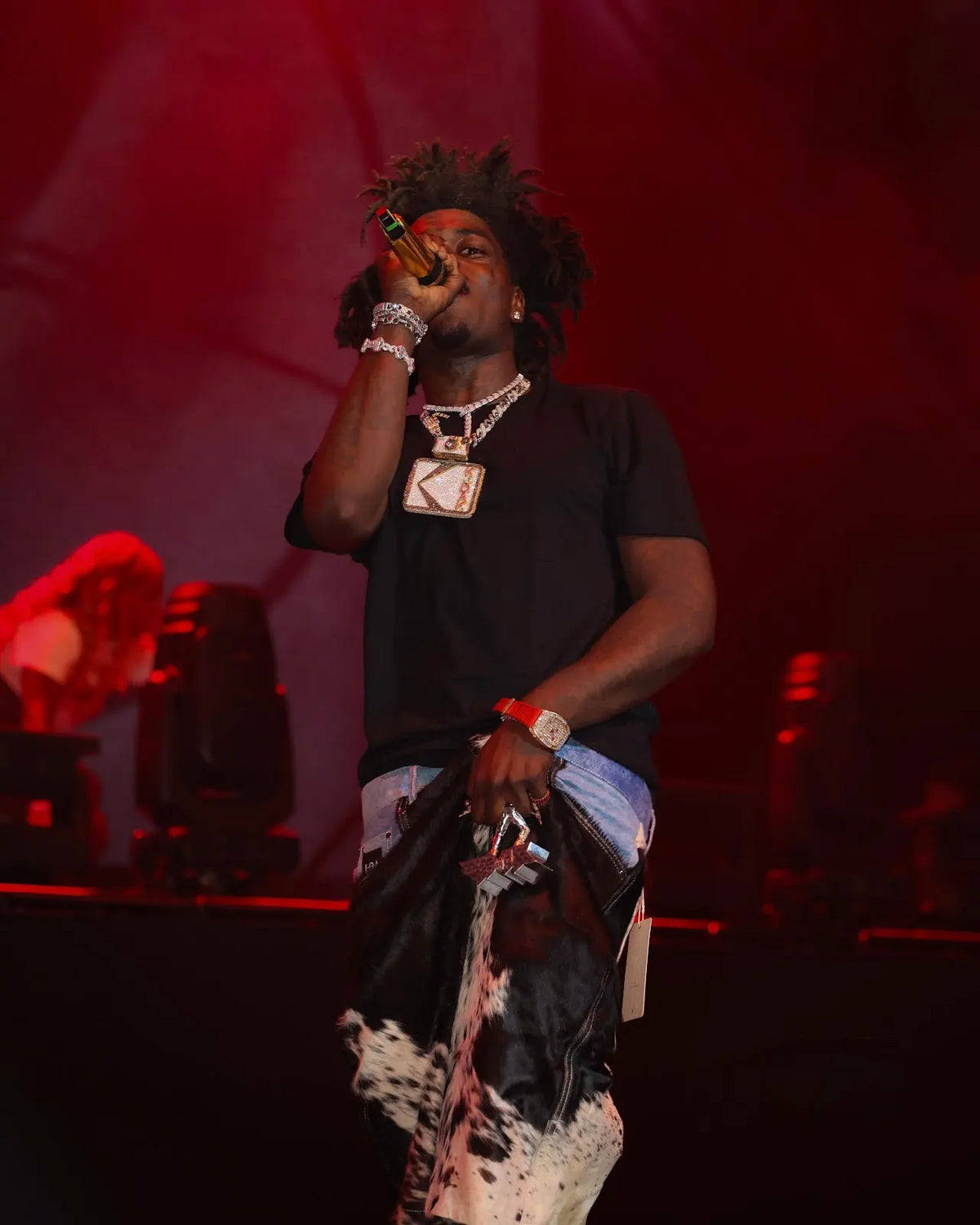 KODACK on stage with a microphone, wearing black-and-white cowhide jorts with a denim waistband.