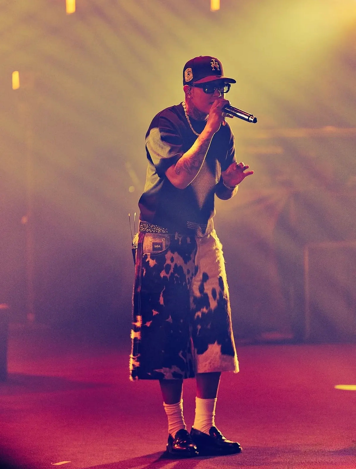 KWN performing under warm stage lights in cowhide jorts, a black T-shirt and a cap.