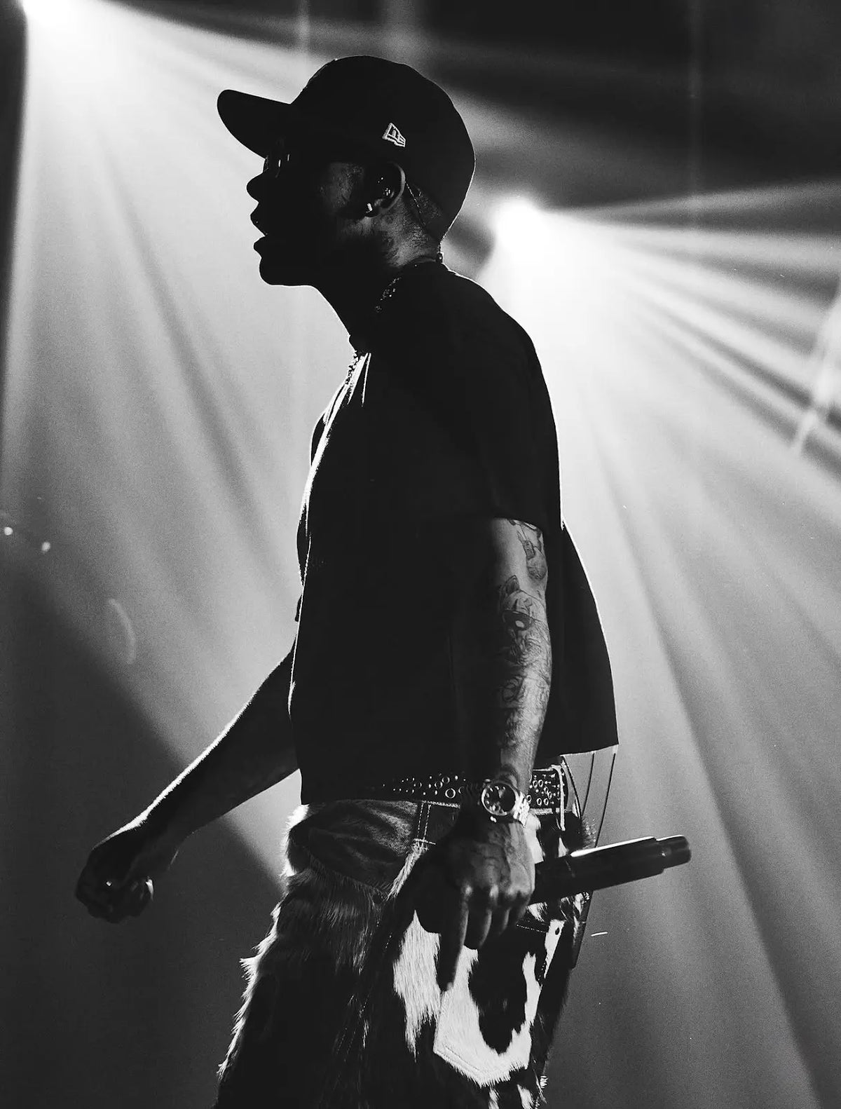 Black-and-white stage photograph of KWN in a cap, T-shirt and cowhide jorts, holding a microphone.