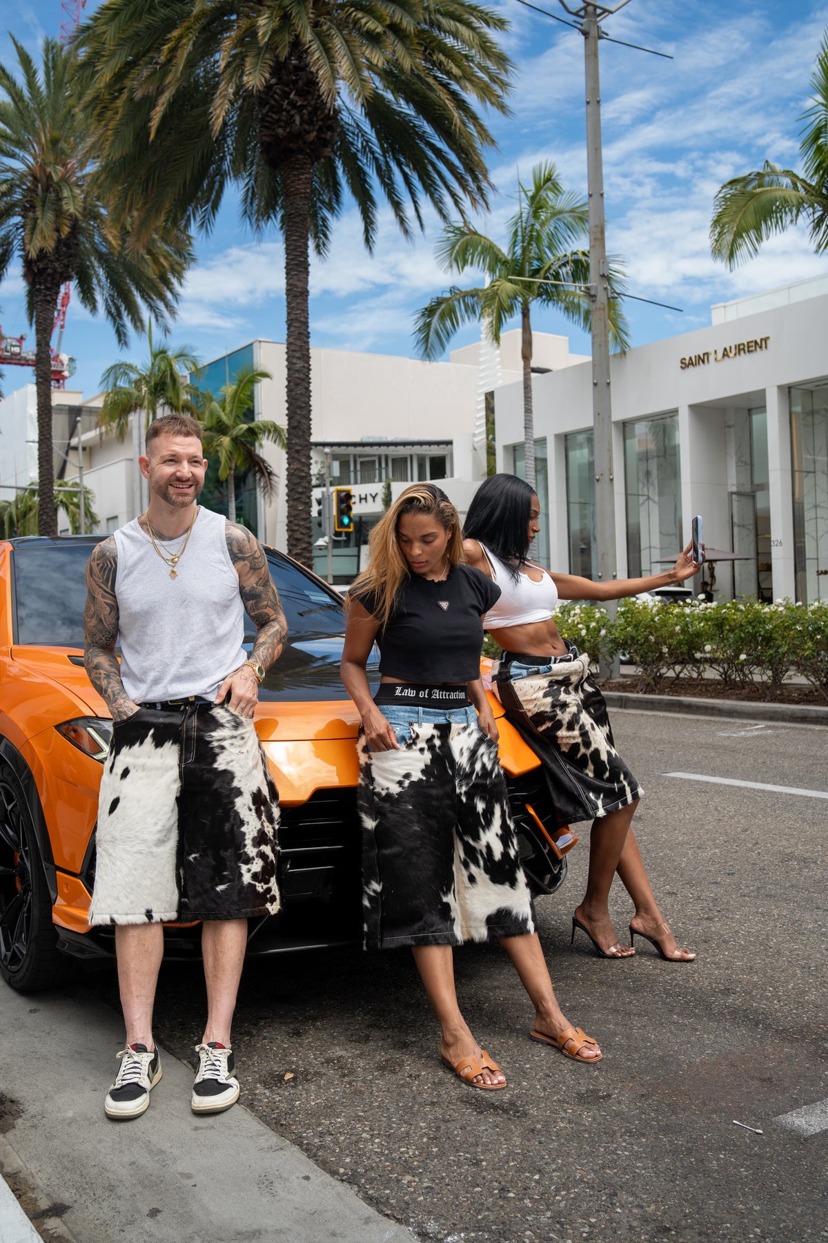 Three people wearing Law of Attraction cowhide denim by an orange car on a palm-lined street
